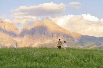 A couple walks across a flowering meadow towards the mountain range at sunset, Alpe di Siusi,