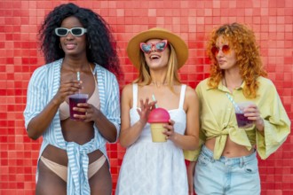 Three diverse women enjoying refreshing drinks against a vibrant red wall, celebrating summer and