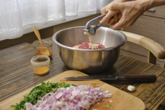 Hands mincing garlic into a metal bowl filled with ground meat while chopped onions and fresh herbs