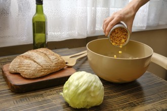 A hand pours canned corn into a mixing bowl on a wooden table. Nearby, a head of lettuce and a loaf