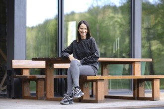 A woman with a bright smile is sitting at a rustic wooden bench on the porch of modern glass cabin,