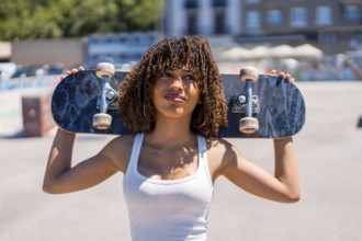 Confident young woman holding skateboard on shoulders, enjoying sunny day at skatepark, showcasing