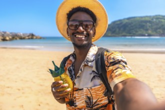 Young black man enjoying summer vacations on a tropical beach, taking a selfie while holding a