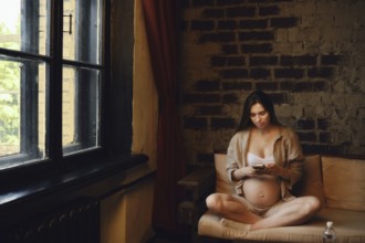 A woman in her third trimester sits comfortably on a sofa at home. She is wearing casual clothing