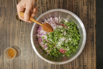 A hand holds a wooden spoon filled with spices over a stainless steel bowl containing chopped