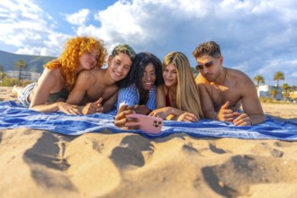 Five cheerful young friends are lying on a beach towel, taking a selfie with a smartphone, enjoying