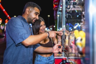 Happy couple enjoying a night out, playing a claw crane game at an amusement park, trying their