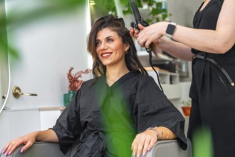 Hairdresser styling hair of a smiling client with a curling iron in a chic, modern beauty salon,