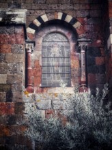 An ornate stained glass window featuring detailed artwork is set into a weathered stone wall.