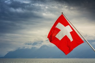 View of the Swiss flag waving over Lake Geneva with mountains and clouds in Switzerland during a