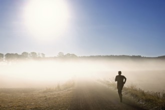 Man training running on a gravel road in the morning fog in a rural landscape with the sun shining