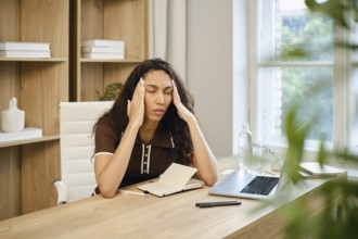 A woman with curly hair sits at a wooden desk in a stylish home office, visibly stressed while