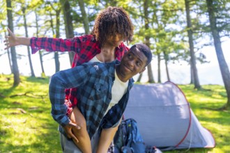 Young man giving piggyback ride to girlfriend in forest near tent, enjoying camping trip in nature