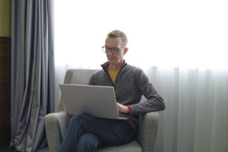 A man wearing glasses sits comfortably in a gray chair and fills out a tax return online on his