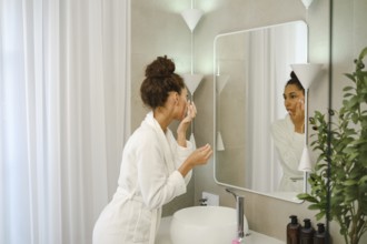 A woman in a white robe is applying skincare in a stylish bathroom. She is focused on her