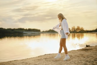 A young woman with long blonde hair walks along the sandy beach during sunset. She is happy to be
