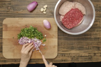 A person chops fresh herbs and onions on a wooden cutting board while ground meat rests in a