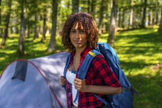 Young woman wearing a backpack enjoys camping in a serene forest, relaxing near her tent and