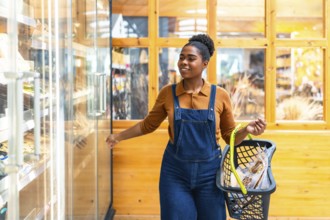 Smiling black woman opening refrigerated section door in grocery store holding shopping basket