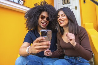 Two friends are sitting on the steps of a vibrant yellow building, laughing and watching a video on