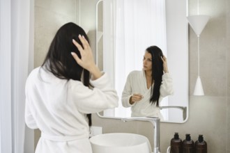 A woman stands in front of a mirror in a modern bathroom, dressed in a white bathrobe. She is