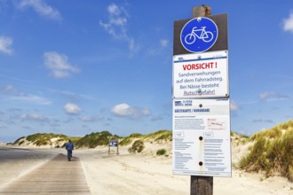 Cyclist on wooden path through the dunes to the sea, sign warns of wetness, danger of slipping and