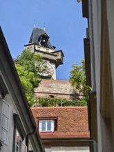 Historic clock tower towers over rooftops, surrounded by green trees and blue sky Graz Clock Tower