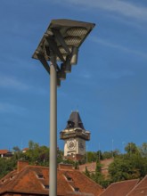 Clock tower behind a street lamp, surrounded by historic roofs and trees Graz Clock Tower Graz