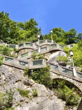 Stone wall with green railings and lamps on a steep, wooded slope under a blue sky Graz Styria