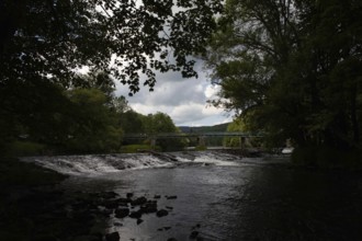 A dark river with a waterfall and bridge, surrounded by forests and cloudy skies Hardegg