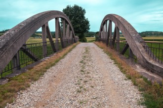 An old bridge with a gravel path leads through a rural area, surrounded by fields and a clear sky