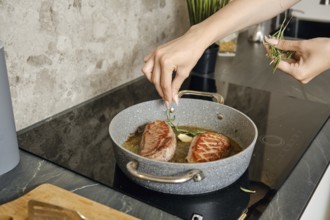A person is preparing two pieces of duck breast in a frying pan, adding fresh herbs to enhance