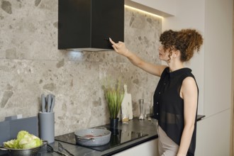 A woman prepares a healthy meal in a stylish kitchen, adjusting the vent fan above her cooking area
