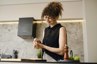 A woman stands in a stylish kitchen, smiling as she seasons a dish with a spice grinder. The