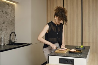 A woman with curly hair is cutting raw meat in a kitchen. Natural light illuminates her workspace