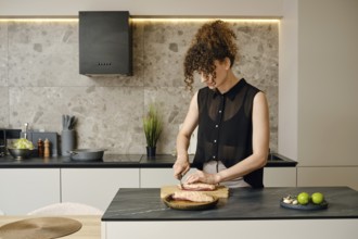 In a sleek, contemporary kitchen, a woman with curly hair skillfully chops ingredients on a wooden