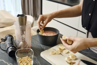A person adds slices of bananas to a creamy smoothie mixture in a black bowl. Nearby, a blender and