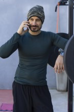 Athlete takes a break from his workout, leaning against a weightlifting rack and talking on his