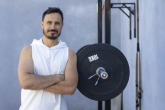 Muscular personal trainer with crossed arms leans confidently against a weight plate in an outdoor