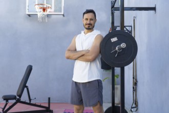 Sportsman with crossed arms is posing next to a weightlifting rack in a private outdoor gym,
