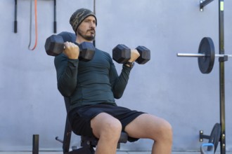 Focused male athlete sitting on a weight bench, lifting dumbbells and performing bicep curls during