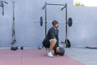 Young sportsman wearing beanie and thermal shirt performing wall ball squats in an outdoor gym,