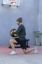 Young sportsman wearing a beanie and sportswear is lunging with a medicine ball in an outdoor gym,