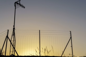 Silhouette of a television antenna and a wire mesh fence against a vibrant sunset sky
