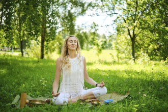 A woman sits cross-legged on a mat in a lush green park, eyes closed and arms resting on her knees.