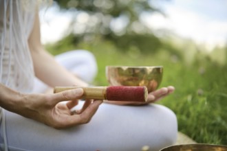 A person is holding a red-tipped mallet next to a singing bowl, seated on grass in a serene outdoor