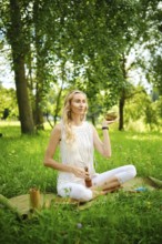 A woman practices meditation outdoors, holding a singing bowl while seated on a mat in a field. The