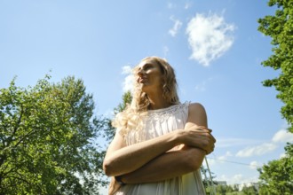In a green park on a bright day, a woman stands with her arms crossed, gazing upwards. Sunlight
