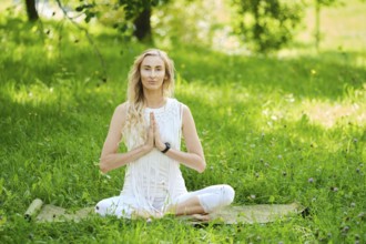 A woman dressed in white sits cross-legged on a mat, practicing yoga outdoors. Surrounded by