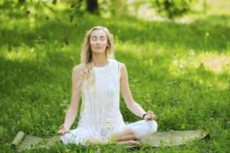 A woman in a white outfit sits cross-legged on a mat, eyes closed and hands resting on her knees in